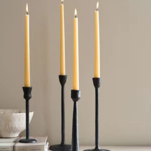 Four tall, slender candles are displayed in black metal candle holders. The candles are a soft yellow color, each with a small flame at the top. The holders vary slightly in height and design, featuring a simple, modern aesthetic. In the foreground, a light-colored bowl sits atop a stack of books, adding a touch of warmth to the arrangement against a neutral background.