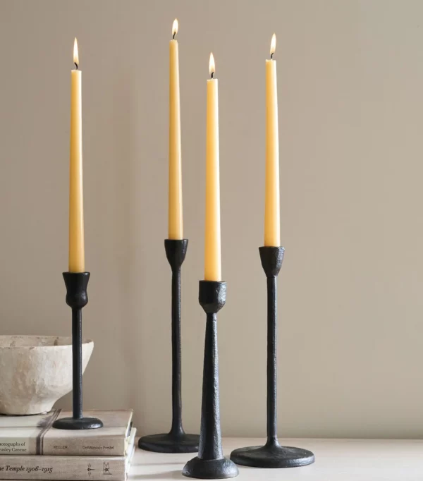 Four tall, slender candles are displayed in black metal candle holders. The candles are a soft yellow color, each with a small flame at the top. The holders vary slightly in height and design, featuring a simple, modern aesthetic. In the foreground, a light-colored bowl sits atop a stack of books, adding a touch of warmth to the arrangement against a neutral background.