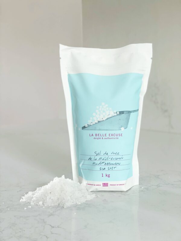A white pouch of sea salt is prominently displayed, featuring a light blue design with an illustration of salt being poured. The label reads "LA BELLE EXCUSE" along with text in French and English indicating it is Mediterranean sea salt, weighing 1 kg. A small mound of salt is spilled on the surface in front of the pouch, which is set against a light, smooth background.