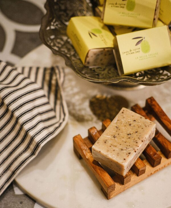 A bar of soap sits on a wooden soap dish, featuring a light beige color with small dark specks. Behind it, a decorative metal bowl holds several wrapped soap bars, each in yellow packaging with a green label. A striped cloth napkin is partially visible to the left, adding texture to the scene. The background consists of a smooth, light-colored surface, enhancing the overall aesthetic.