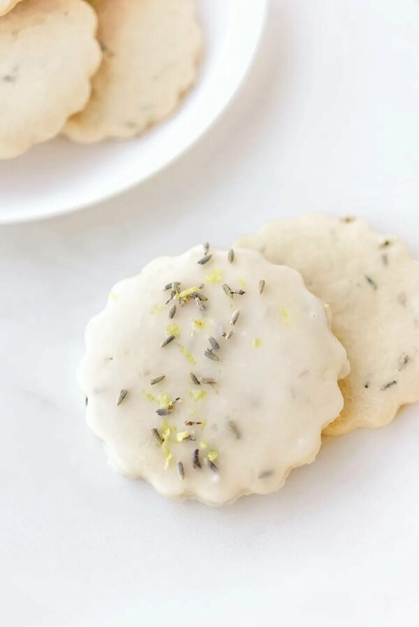 A close-up of round, scalloped cookies is displayed. One cookie is topped with a glossy white icing, sprinkled with dried lavender flowers and small bits of lemon zest. Two additional cookies are partially visible behind it, showcasing a light beige color with hints of lavender. The background is a smooth, light surface, enhancing the delicate appearance of the cookies.