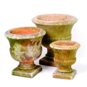 Three weathered terracotta garden urns of varying sizes, each with a square base and a wide, flared rim. The urns show signs of moss and lichen growth, giving them a rustic, aged appearance. The colors range from earthy orange to greenish hues due to the natural patina.