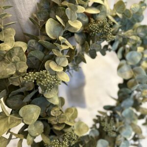 A decorative wreath made of various shades of green eucalyptus leaves and small clusters of berries. The leaves vary in size and shape, creating a lush, textured appearance. The background is softly blurred, emphasizing the wreath's intricate details.