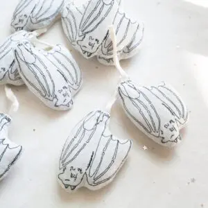 Several small fabric ornaments shaped like bats are shown. They are white with black line drawings depicting bat features, including wings and faces. Each bat has a white ribbon loop attached for hanging. The ornaments are placed on a light-colored surface with some small star-shaped confetti scattered around.