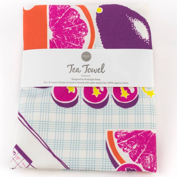 A folded tea towel featuring a vibrant design with colorful fruit illustrations, including citrus slices and star shapes. The towel is predominantly white with a light blue grid pattern in the background. It is labeled with a band that reads "Tea Towel," indicating it is designed by Porchlight Press and made from 100% organic cotton, measuring 29 x 19 inches. The towel is screen-printed using water-based inks.