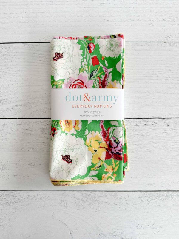 A stack of colorful fabric napkins is displayed, featuring a vibrant floral pattern on a green background. The napkins are neatly folded and secured with a branded paper band that reads "dot & army" along with the text "EVERYDAY NAPKINS" and "made in georgia." The background consists of light wooden planks, enhancing the overall aesthetic.