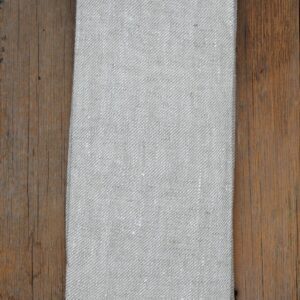 A folded piece of light gray fabric is displayed on a wooden surface. The fabric has a textured weave with subtle variations in color and a frayed edge at the bottom. The wood beneath is a warm brown, adding contrast to the fabric.