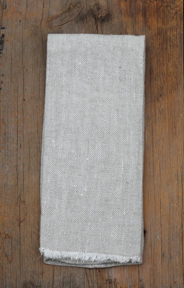 A folded piece of light gray fabric is displayed on a wooden surface. The fabric has a textured weave with subtle variations in color and a frayed edge at the bottom. The wood beneath is a warm brown, adding contrast to the fabric.
