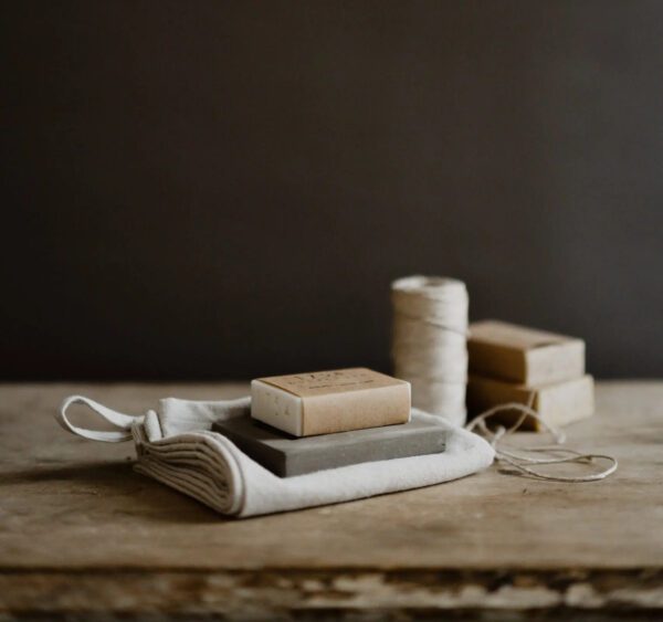 A rustic wooden surface displays a collection of items. At the forefront, two soap bars are stacked on a small, folded cloth pouch. The top soap has a light brown wrapper with text, while the bottom soap is dark gray. Behind them, a spool of twine sits next to several wrapped soap bars, all in neutral tones. The background is a muted dark color, enhancing the earthy aesthetic of the arrangement.
