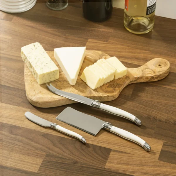 A wooden cutting board displays three types of cheese: a block of herbed cheese, a triangular piece of soft cheese, and several slices of yellow cheese. Accompanying the cheese are three cheese knives with white handles: a curved knife, a flat cheese knife, and a small spreading knife. The setting features a wooden countertop with a few bottles in the background.