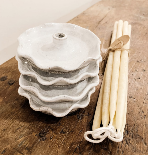 A stack of four white ceramic dishes with a wavy edge is positioned on a wooden surface. The top dish has a small central hole. Next to the dishes, there are several cream-colored taper candles tied together with twine, featuring a small tag. The overall aesthetic is rustic and minimalistic.