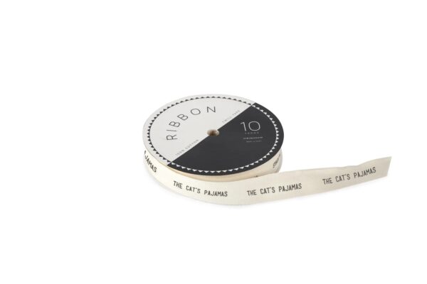A roll of twill ribbon is displayed, featuring a cream color with the phrase "THE CAT'S PAJAMAS" printed in black. The ribbon is made of 100% cotton and measures 10 yards in length. The spool has a black and white design, with a decorative edge on the black section.