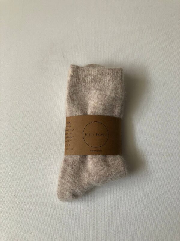 A pair of soft, light beige socks is neatly folded and secured with a brown paper band. The band features a circular logo with the text "Billy Bamboo" and several words printed around it, likely describing the product's qualities. The socks appear fluffy and cozy, suggesting warmth and comfort. The background is a plain white surface, emphasizing the socks and their packaging.
