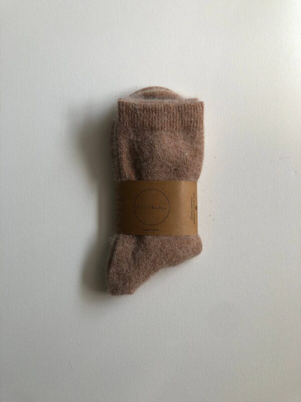 A pair of soft, light brown socks is displayed, partially rolled up. The socks have a fuzzy texture and are secured with a circular, brown paper band that features the brand name "Billy Bamboo." The background is a plain white surface, enhancing the focus on the socks.