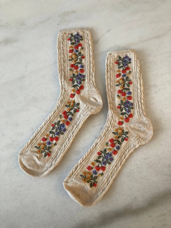 A pair of cream-colored socks featuring intricate floral embroidery along the sides. The design includes red, blue, and yellow flowers intertwined with green leaves. The socks have a textured pattern along the edges, adding a decorative touch. They are laid flat on a light marble surface.