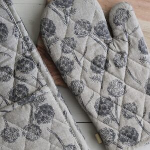 A pair of quilted oven mitts is prominently displayed, featuring a floral pattern with dark gray illustrations of flowers and leaves on a light beige background. The mitts are positioned next to a wooden cutting board, which adds a natural element to the scene. The overall aesthetic is rustic and cozy, suitable for a kitchen setting.