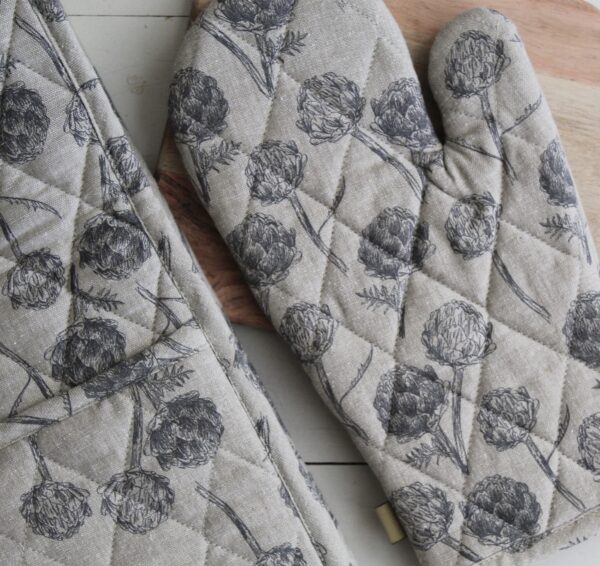 A pair of quilted oven mitts is prominently displayed, featuring a floral pattern with dark gray illustrations of flowers and leaves on a light beige background. The mitts are positioned next to a wooden cutting board, which adds a natural element to the scene. The overall aesthetic is rustic and cozy, suitable for a kitchen setting.