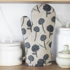 A quilted oven mitt with a floral design featuring dark blue flowers and green stems is prominently displayed. It has a textured, beige background and is positioned next to a white ceramic container with a lid and a smaller green ceramic dish. The setting appears to be a kitchen with a marble backdrop.