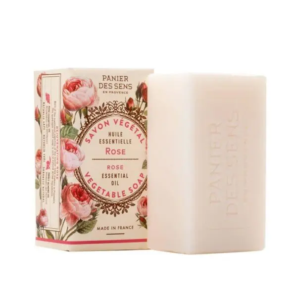 A rectangular bar of soap is displayed next to its decorative packaging. The soap is white and features a subtle embossed design. The packaging is adorned with illustrations of pink roses and includes text in both French and English, indicating it is a vegetable soap with rose essential oil. The brand name, "Panier des Sens," is prominently featured on both the box and the soap itself. The overall aesthetic is elegant and floral, suggesting a luxurious bathing experience.