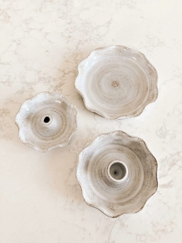 Three ceramic dishes are arranged on a light-colored, marbled surface. The dishes vary in size, with the largest on the right featuring a scalloped edge and a central hole. The middle dish is slightly smaller, also with a scalloped design, while the smallest dish on the left has a similar shape and a single hole in the center. All dishes have a soft, muted color with subtle speckles, giving them a rustic appearance.