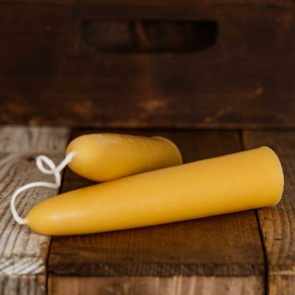 Two yellow beeswax candles are positioned on a wooden surface. The candles have a smooth, cylindrical shape, tapering to a point at one end. One candle is larger and longer, while the other is shorter and rounder. A white wick is attached to the shorter candle, adding a decorative touch. The background features a rustic wooden texture, enhancing the natural aesthetic of the scene.