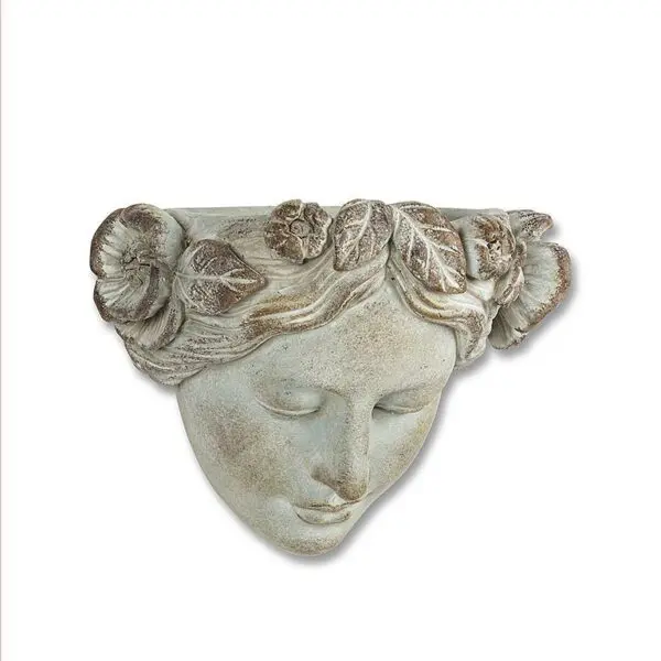 A decorative wall sculpture depicting a serene female face. The figure features a floral crown made of roses and leaves, intricately detailed. The surface has a weathered, stone-like finish with soft, muted colors, giving it an antique appearance. The expression is calm and peaceful, with closed eyes and a slight smile.