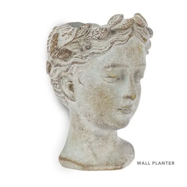 A decorative wall planter shaped like a classical bust of a young woman. The planter features a textured surface with a weathered finish, showcasing intricate details such as a crown of leaves adorning her head. The overall color is a soft, muted tone with hints of green and brown, giving it an antique appearance. The planter is designed to hold plants or flowers, with an opening at the top for planting.