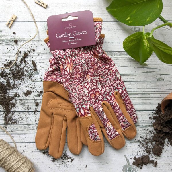 A pair of garden gloves is displayed, featuring a floral pattern in shades of red and cream on the fabric portion and a tan leather-like material on the palms and fingers. The gloves are partially folded, revealing the design and texture. A tag labeled "Garden Gloves" is attached, indicating the brand "Orange Grove" and mentioning "William Morris Collection." Surrounding the gloves are small piles of dark soil and a length of twine, with a green plant visible in the background. The surface beneath is light-colored wood.