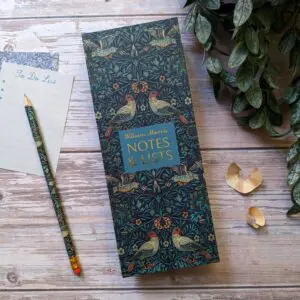 A decorative notepad titled "William Morris NOTES & LISTS" features an intricate design with birds and foliage in rich colors. Next to it, there is a light-colored "To Do List" notepad with lines and a pencil adorned with a matching pattern. Two small gold paper clips are placed nearby, and a green leafy plant is partially visible in the background. The setting has a warm, natural wood texture.