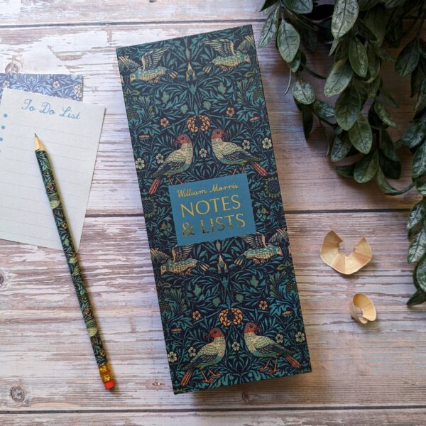 A decorative notepad titled "William Morris NOTES & LISTS" features an intricate design with birds and foliage in rich colors. Next to it, there is a light-colored "To Do List" notepad with lines and a pencil adorned with a matching pattern. Two small gold paper clips are placed nearby, and a green leafy plant is partially visible in the background. The setting has a warm, natural wood texture.
