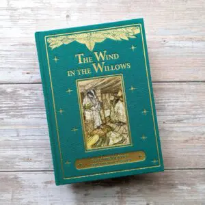 A hardcover edition of "The Wind in the Willows" is displayed. The book features a teal cover with gold lettering and decorative elements. An illustration is centered on the front, depicting two characters in a rustic setting surrounded by various vegetables. The bottom of the cover includes the text "Balth Treasury of Children's Classics." The background is a light wooden surface, enhancing the book's classic aesthetic.
