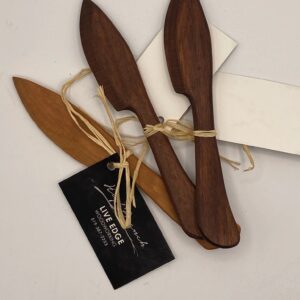 A collection of wooden knives is arranged on a light surface. There are three dark wooden knives with pointed tips and smooth edges, and one lighter wooden knife. They are tied together with natural twine. A small black tag with white text is attached, indicating "LIVE EDGE WOODWORKING" along with a phone number. A plain white card is also included in the arrangement.