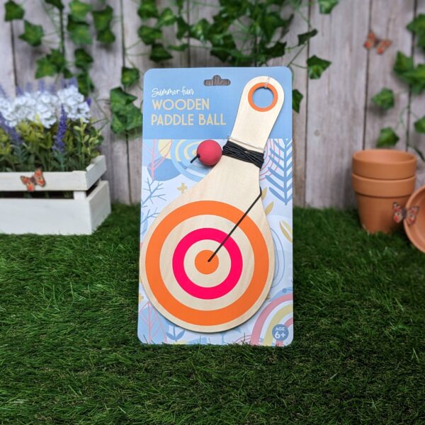 A wooden paddle ball toy is prominently displayed, featuring a circular target design in orange and pink. The paddle has a red ball attached by a black elastic string. The packaging includes colorful graphics and the text "Summer Fun Wooden Paddle Ball," indicating it's suitable for ages 6 and up. In the background, there are potted plants and terracotta pots, adding a natural touch to the setting. The surface is covered with artificial grass.