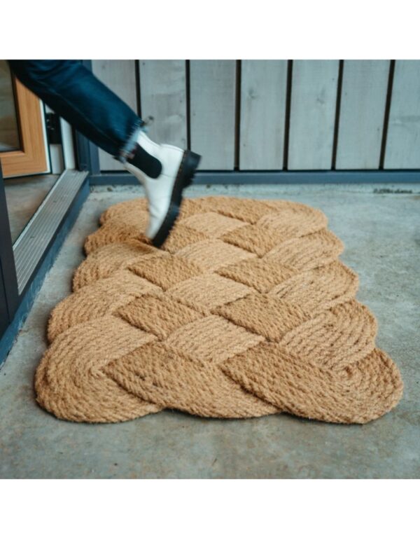 A textured, braided doormat in a warm beige color is positioned at the entrance. The mat features a distinctive woven pattern that resembles a braid, adding a decorative touch. A foot in a white boot with a black elastic side is stepping onto the mat, suggesting a welcoming entryway. The background includes a wooden door and a wall with vertical wooden panels.