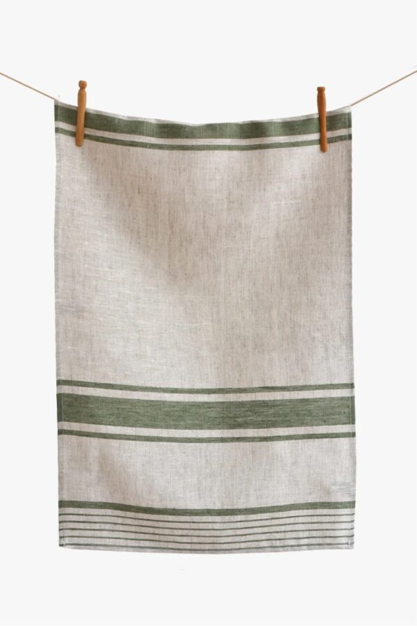 A textured linen towel is hanging from a wooden peg on a string. The towel features a light beige base with two horizontal green stripes at the top and a series of thin green lines towards the bottom, creating a simple yet elegant design. The fabric appears soft and natural, emphasizing a rustic aesthetic.
