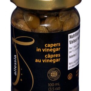 A glass jar with a gold lid contains capers in vinegar. The jar is labeled with the text "capers in vinegar" and "câpres au vinegar" in yellow font on a black background. The jar holds 100 mL (3.5 oz) of capers, which are visible inside. Nutritional information is printed on the side of the jar.