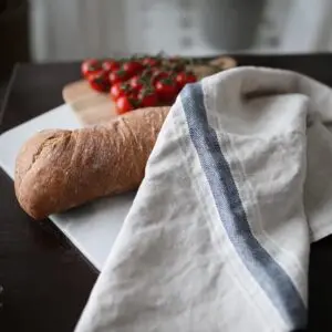 A rustic loaf of bread rests on a white cutting board, accompanied by a cluster of vibrant cherry tomatoes. A light-colored linen cloth with a dark blue stripe is draped nearby, adding a touch of texture to the scene. The background features soft, natural light filtering through sheer curtains, enhancing the warm, inviting atmosphere.