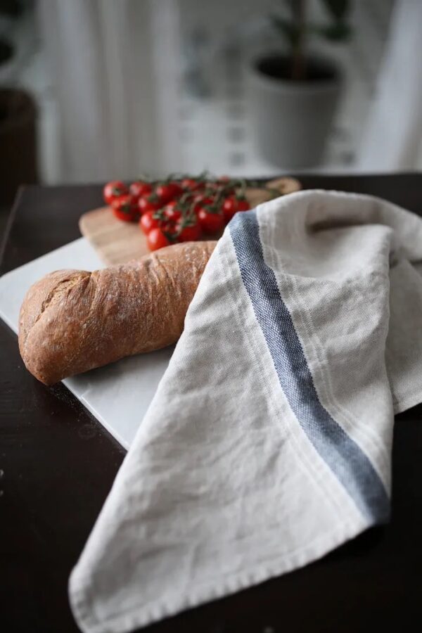 A rustic loaf of bread rests on a white cutting board, accompanied by a cluster of vibrant cherry tomatoes. A light-colored linen cloth with a dark blue stripe is draped nearby, adding a touch of texture to the scene. The background features soft, natural light filtering through sheer curtains, enhancing the warm, inviting atmosphere.