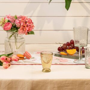 A beautifully arranged table setting features a variety of elements. In the center, a clear glass vase holds pink and orange flowers, complemented by a floral table runner. To the left, pink tulips and colorful macarons (orange and pink) are placed on the table. A yellow-tinted glass sits prominently in the foreground, while two additional glasses, one clear and one textured, are positioned to the right. A small tray holds fresh grapes and a sliced orange, adding a vibrant touch to the scene. The background consists of light wooden panels, enhancing the overall aesthetic.
