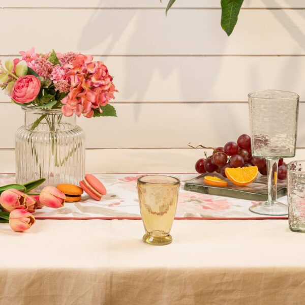 A beautifully arranged table setting features a variety of elements. In the center, a clear glass vase holds pink and orange flowers, complemented by a floral table runner. To the left, pink tulips and colorful macarons (orange and pink) are placed on the table. A yellow-tinted glass sits prominently in the foreground, while two additional glasses, one clear and one textured, are positioned to the right. A small tray holds fresh grapes and a sliced orange, adding a vibrant touch to the scene. The background consists of light wooden panels, enhancing the overall aesthetic.