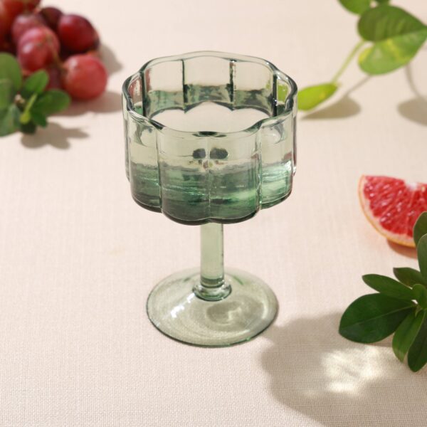 A green glass dessert cup with a scalloped edge sits on a light-colored tablecloth. The cup has a sturdy stem and a wide bowl, partially filled with liquid. Surrounding the cup are a cluster of red grapes, a slice of grapefruit, and some green leaves, adding a fresh and vibrant touch to the scene.