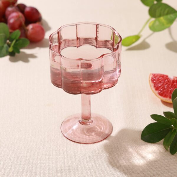 A pink glass dessert cup with a scalloped edge sits on a light-colored textured surface. The cup has a short stem and a wide base, creating an elegant silhouette. Nearby, there are clusters of red grapes, a slice of grapefruit, and some green leaves, adding a fresh and vibrant touch to the scene. The overall composition is bright and inviting.