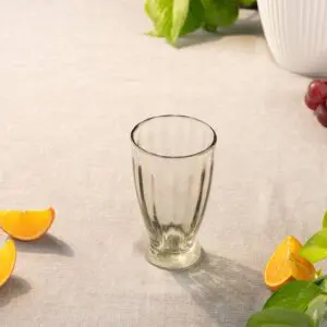 A clear glass tumbler is positioned upright on a light-colored linen tablecloth. Surrounding the glass are several orange segments and a few green leaves, adding a fresh, vibrant touch to the scene. In the background, a white ribbed vase can be seen, along with some grapes. The overall composition conveys a casual, inviting atmosphere.