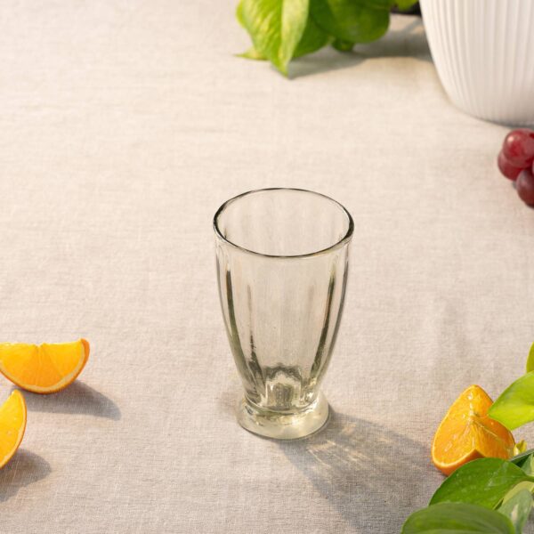 A clear glass tumbler is positioned upright on a light-colored linen tablecloth. Surrounding the glass are several orange segments and a few green leaves, adding a fresh, vibrant touch to the scene. In the background, a white ribbed vase can be seen, along with some grapes. The overall composition conveys a casual, inviting atmosphere.