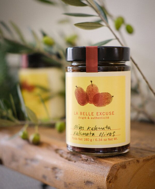 A glass jar with a black lid is prominently displayed, featuring a yellow label adorned with an illustration of two olives. The label reads "LA BELLE EXCUSE" along with the text "olives kalamata" in a handwritten style. The jar contains 180 grams (6.34 oz) of kalamata olives. It is placed on a rustic wooden surface, surrounded by olive branches, creating a natural and inviting atmosphere.