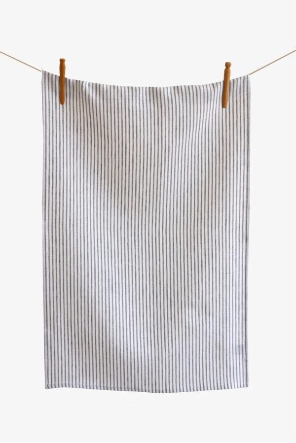 A striped fabric towel is hanging on a clothesline, secured with wooden clips. The towel features a pattern of thin, vertical stripes in light and dark shades, creating a simple yet elegant design. The background is neutral, emphasizing the towel's texture and pattern.