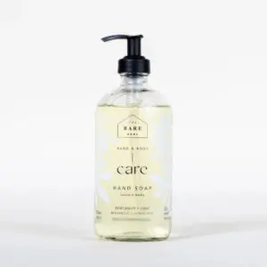 A clear glass bottle of hand soap with a black pump dispenser. The label features a minimalist design with the text "THE BARE HOME," "HAND & BODY," and "care HAND SOAP" prominently displayed. The soap is a light yellow color, and the label includes decorative white leaf patterns. It is scented with "Bergamot + Lime," as indicated on the label. The bottle holds 476 mL (16 fl oz) of product.