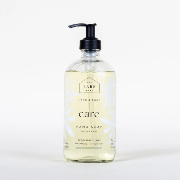 A clear glass bottle of hand soap with a black pump dispenser. The label features a minimalist design with the text "THE BARE HOME," "HAND & BODY," and "care HAND SOAP" prominently displayed. The soap is a light yellow color, and the label includes decorative white leaf patterns. It is scented with "Bergamot + Lime," as indicated on the label. The bottle holds 476 mL (16 fl oz) of product.