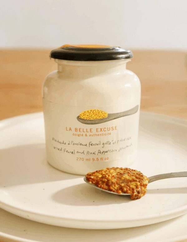 A ceramic jar with a black lid contains a mustard labeled "LA BELLE EXCUSE." The jar features a graphic of a spoon with mustard seeds and text in both French and English, indicating it contains wild fennel and pink peppercorn mustard. A silver spoon rests beside the jar, holding a generous amount of the mustard, showcasing its grainy texture and yellow-brown color. The setting includes a simple white plate and a soft, neutral background.