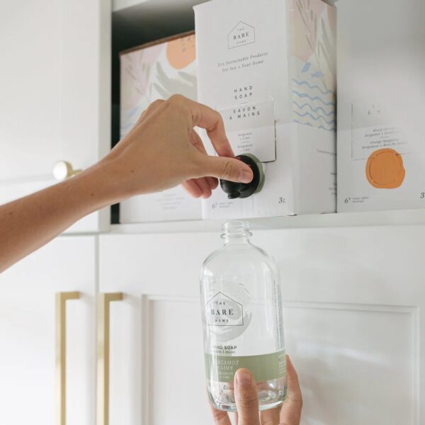 A hand is reaching for a black dispensing nozzle on a box labeled "Hand Soap" from "The Bare Home." Below, a clear glass bottle with a green label reading "Bergamot Lime" is being held, ready to be filled. The background features a white cabinet with gold handles and another decorative box partially visible. The setting appears clean and organized, emphasizing eco-friendly products.